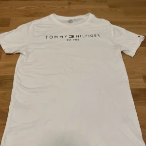 Vit t-shirt från Tommy Hilfiger - Vit t-shirt från Tommy Hilfiger med tryckt logga och text på bröstet. Klassisk rund halsringning och korta ärmar. Perfekt basic-plagg med stilren design.