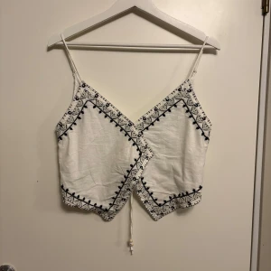 Vit broderad linnetopp från Zara - Superfin vit linnetopp från Zara med svarta broderade detaljer och pärlor. Toppen har smala axelband, omlott-design och knyt i midjan. Perfekt för sommaren och ger en bohemisk känsla med sina unika mönster och detaljer.