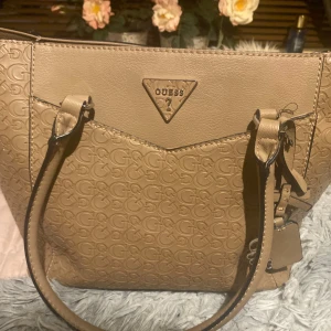 Beige handväska från Guess - Säljer en beige handväska från Guess med präglat logomönster över hela väskan. Väskan har två handtag, metallogo framtill och innerfack med dragkedja. Perfekt för dig som gillar stilrena accessoarer med tydlig logga.