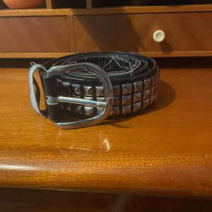Nitbälte med silverdetaljer (RESERVERAD) - Säljer ett svart bälte med tre rader silverfärgade pyramidnitar och ett silverspänne. Bältet har en klassisk punkstil och passar perfekt till jeans eller kjol för en edgy look.