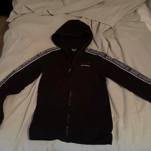 Hoodie - Svart hoodiejacka med dragkedja från EA7 Emporio Armani. Jackan har huva och vita logoband längs ärmarna. Perfekt för en sportig och avslappnad stil. Storlek 160