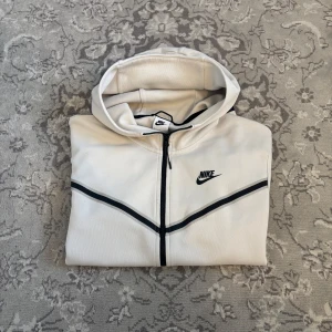 Vit hoodiejacka från Nike med svarta detaljer - Säljer en vit Nike tech från Nike med svarta dragkedjor och detaljer. Tröjan har huva, lång ärm och en ficka med dragkedja på ena ärmen. Perfekt för en sportig och stilren look.
