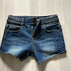 Mörkblå korta jeansshorts med flätat bälte från Dollhouse - Snygga mörkblå korta jeansshorts från Dollhouse med flätat bälte i midjan och dubbla knappar framtill. Klassisk femficksmodell med lätt slitningar och uppvikta benslut. Perfekta för sommaren! Midjemått ca 38 cm