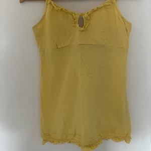 Gul linnetopp från 365 Sunshine - Säljer en söt gul linnetopp från 365 Sunshine i storlek S. Toppen har smala axelband, liten volangdetalj och en dekorativ öppning framtill. Perfekt för varma dagar och enkel att matcha med jeans eller kjol.