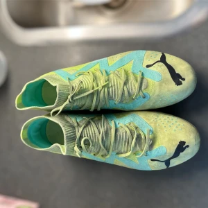 Puma fotbollsskor i limegrönt och turkos - Säljer ett par limegröna och turkosa Puma fotbollsskor med svarta Puma-loggor. Skorna har snörning och mönstrad ovandel samt dobbar för spel på gräs. Insidan är turkos och ovandelen har en stickad känsla för extra komfort.