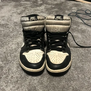 Nike Air Jordan 1 Retro High OG mocha  - Säljer ett par Nike Air Jordan 1 Retro High OG i svart och vitt med högt skaft och klassisk Nike-logga på sidan. Skorna har snörning och perforerad tå för extra ventilation. Perfekta för dig som gillar streetwear och sneakers med ikonisk design.