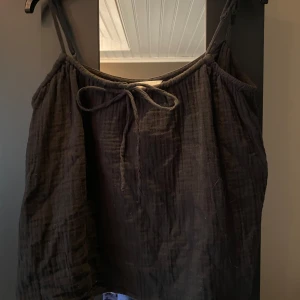 Svart linnetopp med knyt från H&M - Svart linnetopp från H&M med tunna axelband och knytning framtill. Toppen har en luftig och lätt struktur, perfekt för varma dagar. Enkel och stilren design som passar till mycket.