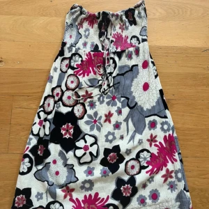 Blommig tubklänning från Vero Moda - Har knutit bandet som halterneck, då jag inte har byst att hålla upp klänningen! Lite lös i resåren högst upp i i tuben! Kan användas som kjol! Längd 79cm! Säljer en blommig tubklänning från Vero Moda i storlek M. Klänningen har ett färgglatt mönster i svart, grått, vitt och rosa med smock upptill och knytning framtill. Perfekt för varma dagar och har en mjuk bomullskänsla.