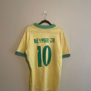 Brasilien Neymar Jr 10 matchtröja - Gul Brasilien fotbollströja med gröna detaljer, Neymar Jr och nummer 10 på ryggen. Officiell Nike-tröja med CBF-märke och Brasil-tryck på bröstet. Kortärmad med unikt mönster i tyget och gröna muddar vid ärmar och krage.