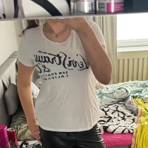 Vit t-shirt från Levis med tryck - Vit t-shirt från Levis  med svart tryck över bröstet. T-shirten har rund halsringning och korta ärmar. Perfekt för en avslappnad stil och enkel att matcha med olika outfits.