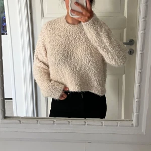 Beige fluffig stickad tröja - Mysig och fluffig beige stickad tröja med rund hals och lång ärm. Tröjan har en något croppad passform och är perfekt för kyligare dagar. Enkel och stilren design som passar till det mesta.