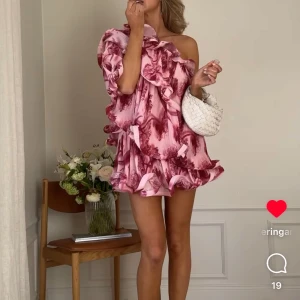 Rosa volangklänning med one shoulder lojsanXnakd - Kikar intresset för  en superfin rosa kortklänning med volanger och one shoulder-design. Klänningen har ett blommigt mönster och är perfekt för dig som vill sticka ut. Den har en luftig passform och är riktigt snygg till sommarens alla tillfällen. Den är helt oanvänd och har original förpackning! Klänningen är helt slutsåld 