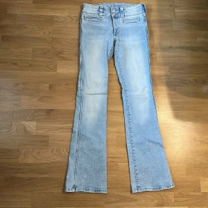 Ljusblå bootcut jeans från H&M - Säljer ett par ljusblå bootcut jeans från H&M med låg midja och justerbar midja. Jeansen har två dekorativa fickor fram och stängs med dubbla knappar och dragkedja. Perfekta för dig som gillar en klassisk jeanslook.