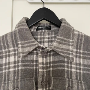 Grårutig overshirt från Manière De Voir - Säljer en grå och vit-rutig overshirt från Manière De Voir i storlek XS. Skjortan har långa ärmar, klassisk krage och knäppning framtill samt två bröstfickor. Perfekt att slänga över en t-shirt för en avslappnad look. Mått: 52cm (armhåla ner), 45 cm (bröstvidd), 72cm (axel ner).