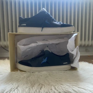 Svarta sneakers från Axel Arigato med blå fågel - Snygga svarta sneakers från Axel Arigato med broderad blå fågel på sidan och vit sula. Skorna har snörning och är tillverkade i skinn. Levereras med originalkartong och dustbag. Skriv för mer bilder och information 