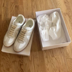 Maison Margiela sneakers i vitt och beige - Säljer ett par stilrena Maison Margiela sneakers i vitt och beige med klassisk gummisula. Skorna har snörning och är tillverkade i en mix av skinn och mocka. Perfekta för dig som gillar minimalistisk och tidlös design.