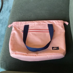 Rosa axelväska från Nike - Säljer en ljusrosa axelväska från Nike med marinblåa handtag och en rödvit dragkedja framtill. Väskan har en enkel, sportig design och är perfekt för att bära med sig det viktigaste. Nike-loggan sitter på en blå lapp i nedre hörnet.
