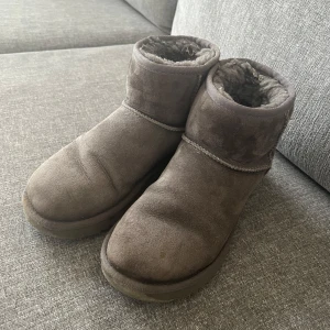 Gråa UGGs i mocka - Mysiga gråa UGGs i mjuk mocka med varmt foder inuti. Skorna har en klassisk rund tå och platt sula, perfekta för kyliga dagar. Utsidan är i mocka och insidan är fodrad med mjuk päls för extra komfort.
