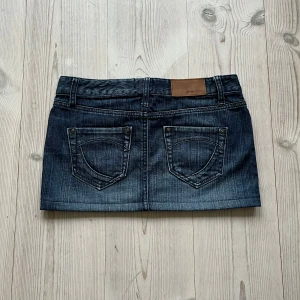 Blå lågmidjad jeanskjol  - Mörkblå lågmidjad jeanskjol från Gina i storlek 36. Fint skock. Tvärs över midjan är det 39 cm och den är ca 27 cm lång. Använd gärna köp nu!