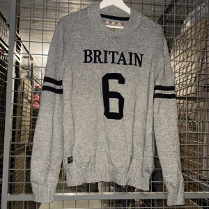 Grå stickad tröja med text från Morris - Säljer en grå stickad tröja från Morris med texten 'BRITAIN' och siffran 6 framtill. Tröjan har svarta ränder på ärmarna och ribbade muddar vid hals, ärmslut och nederkant. Perfekt för dig som gillar sportig stil.