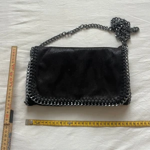 Svart axelväska med kedjedetaljer - Snygg svart axelväska med silverfärgad kedja som axelrem och runt kanterna. Väskan har magnetlås och ett stort huvudfack med innerficka med dragkedja. Perfekt för att bära det viktigaste och ger en cool touch till outfiten.