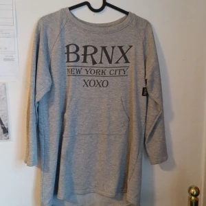 Grå sweatshirt med tryck från Red & Blue - Säljer en grå sweatshirt från Red & Blue med texten 'BRNX NEW YORK CITY XOXO' framtill. Tröjan har långa ärmar och en avslappnad passform. Perfekt för en cool och bekväm stil. ❤️