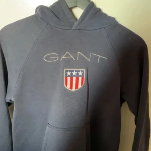 Mörkblå hoodie från GANT - Säljer en mörkblå hoodie från GANT med broderad logga och amerikanskt sköldmärke på bröstet. Tröjan har huva och en stor magficka framtill. Perfekt för en avslappnad stil. Tröjan är i nyskick och jättefin verkligen, ser ny ut, använder inte den och det är dörför jag säljer den