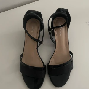 Svarta klackar - Stilrena svarta klackar med öppen tå och ankelrem. Skorna har en platt sula och bred rem över tårna samt justerbar rem runt vristen. Perfekta för varmare dagar och passar till många olika outfits.
