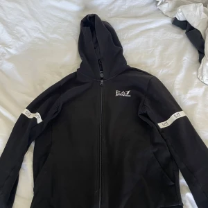 Svart kofta från Emporio Armani EA7 - Svart kofta från Emporio Armani EA7 med dragkedja och huva. EA7-logga på bröstet och vita detaljer med logga på ärmarna. Perfekt för en sportig och stilren look.