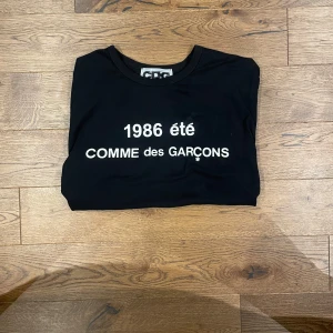 Svart t-shirt från Comme des Garçons - Svart t-shirt med vit text '1986 été COMME des GARÇONS' på bröstet. Klassisk rund halsringning och enkel design som passar perfekt. Fråga gärna efter mer bilder. Använd en gång och har fortfarande kvar necktags om det eftersökes.