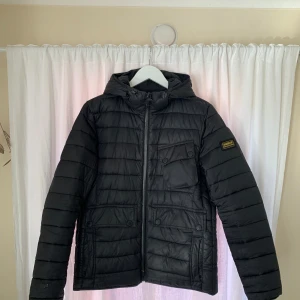   Barbour International - M - Svart quiltad jacka från Barbour International med huva och dragkedja framtill. Jackan har flera fickor, varav en med knapp på bröstet, samt Barbour-logga på ärmen. Perfekt för kyliga dagar.