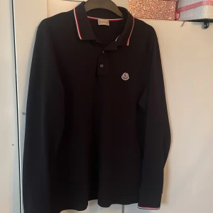 Svart långärmad pikétröja från Moncler - Säljer en svart långärmad pikétröja från Moncler med klassisk krage och knäppning framtill. Tröjan har Moncler-logga på bröstet och rödvit rand på krage och ärmslut. Perfekt för en stilren och sportig look.
