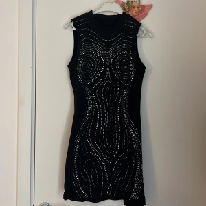 Svart ärmlös klänning med silvriga nitar - Säljer en svart, tight och ärmlös klänning dekorerad med silvriga nitar i ett mönster över hela framsidan. Klänningen har en kroppsnära passform och är kort i modellen. Perfekt för dig som vill sticka ut.