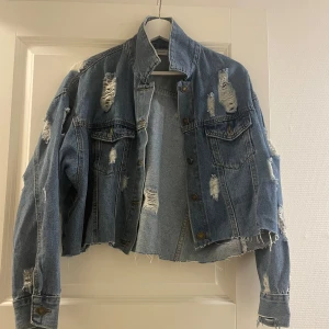 Croppad jeansjacka med slitningar - Säljer en croppad blå jeansjacka med slitna detaljer och rå kant nertill. Jackan har klassisk krage, bröstfickor med lock och knappar framtill. 