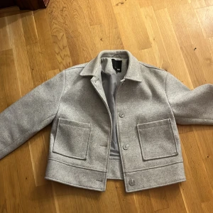 Ljusgrå croppad jacka från lager 157 - Säljer en stilren ljusgrå jacka från lager 157 med croppad passform. Jackan har stora fickor framtill, klassisk krage och knäppning med knappar. Perfekt för dig som gillar minimalistisk stil.