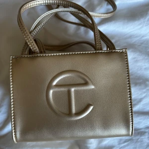 Guldig axelväska från Telfar - Säljer en snygg guldig axelväska från Telfar med präglad logga på framsidan. Väskan har både korta handtag och lång axelrem, perfekt för att bära på olika sätt. Väskan har smink fläckar på insidan som ni kan se på bild, går att få bort i tvätt. Inga andra defekter och använd endast fåtal gånger. Slutsåld online och köpt för orginalpris 