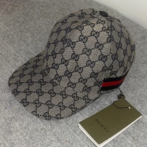 Gucci keps  - För video skicka dm. Snygg keps från Gucci med klassiskt GG-monogram i grått och svart. Kepsen har en röd och svart rand på sidan och justerbar baksida. Märkets logga syns tydligt i mönstret. Perfekt accessoar för en trendig look. Storlek ”M”