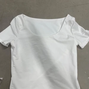 Vit topp med bred urringning - En stilren vit topp med bred, rundad urringning och korta ärmar. Tillverkad i mjuk och stretchig polyesterblandning som sitter bekvämt på kroppen. Perfekt att matcha med jeans eller kjol för en enkel och fräsch look.