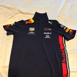 Red Bull Racing t-shirt från Puma - Säljer en mörkblå Red Bull Racing t-shirt från Puma med flera tryckta loggor från sponsorer som Mobil 1, Honda och Pirelli. T-shirten har krage, knappar och korta ärmar. Perfekt för motorsportfans! Köpt i Monaco!!