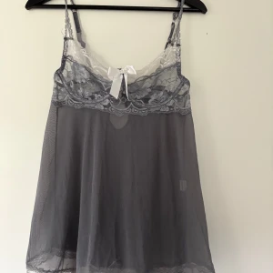 Grå spetslinnetopp med rosett - Säljer en grå linnetopp i mesh med spetsdetaljer och vit rosett framtill. Toppen har tunna axelband, spetskant upptill och dekorativt spänne i ryggen. Nederkanten är dekorerad med spets och ett band som man kan justera. Aldrig använd bara testad 