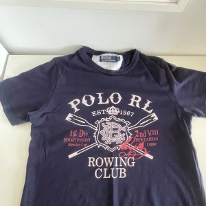 Mörkblå t-shirt från Polo Ralph Lauren - Säljer en mörkblå t-shirt från Polo Ralph Lauren med vit och röd tryckt text och grafik på bröstet. Klassisk rund hals och korta ärmar. Perfekt för en sportig och avslappnad stil.