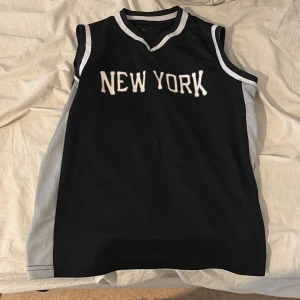 Svart basketlinne med texten NEW YORK från Oxide - Säljer ett svart basketlinne från Oxide med vita och grå detaljer samt texten NEW YORK på bröstet. Linnet har kontrastfärgade kanter och ärmlös design, perfekt för sport eller träning.