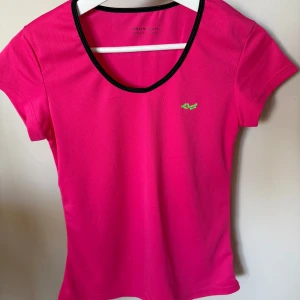 Rosa träningst-shirt från Röhnisch - Snygg rosa t-shirt från Röhnisch med svart kant runt halsen och liten grön logga på bröstet. Perfekt för träning, tillverkad i ett lätt och syntetiskt material som andas. Kortärmad och figurnära passform.