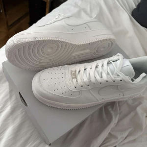 Nike Air Force 1 vita sneakers - Klassiska helvita Nike Air Force 1 sneakers med perforerad tå och snörning. Skorna har en robust sula och är tillverkade i skinn med diskreta Nike-loggor på sidorna. Perfekta för en clean och stilren look.