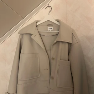 Beige kappa från Kappahl - Stilren beige kappa från Kappahl med stora framfickor och klassisk krage. Jackan har knäppning framtill och en rak, något oversized passform. Perfekt för dig som gillar minimalistisk stil.