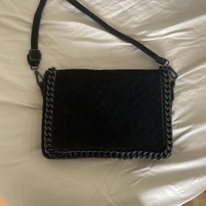 Svart axelväska med kedjedetalj från ALDO - Säljer en svart axelväska från ALDO med snygg kedjedetalj runt kanterna. Väskan har justerbar axelrem och flera fack inuti för bra organisering. Perfekt för dig som vill ha en stilren och trendig accessoar.
