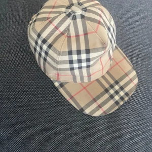 Rutig keps från Burberry - Snygg keps från Burberry i klassiskt beige rutigt mönster med svarta, vita och röda linjer. Kepsen har böjd skärm och justerbar rem baktill. Tillverkad i bomull och polyester för en bekväm passform.
