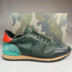 Valentino Rockrunner sneakers i camouflage - Säljer ett par Valentino Rockrunner sneakers i grönt camouflage-mönster med svarta och turkosa detaljer samt röd häl. Skorna har snörning, rund tå och platt sula. Märkeslogga på plösen och nitar på hälen ger en unik touch.