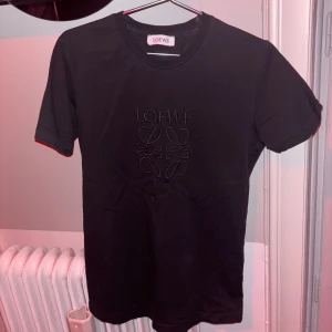 Loewe T-shirt  - Säljer en svart t-shirt från Loewe med broderad Loewe-logga framtill. T-shirten har rund halsringning och korta ärmar. Perfekt för en clean och stilren look.