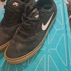 Svarta Nike SB sneakers med vit swoosh - Säljer ett par svarta Nike SB sneakers med vit swoosh och gummisula. Skorna har snörning och en klassisk rund tå. Perfekta för skate eller vardagsstil.